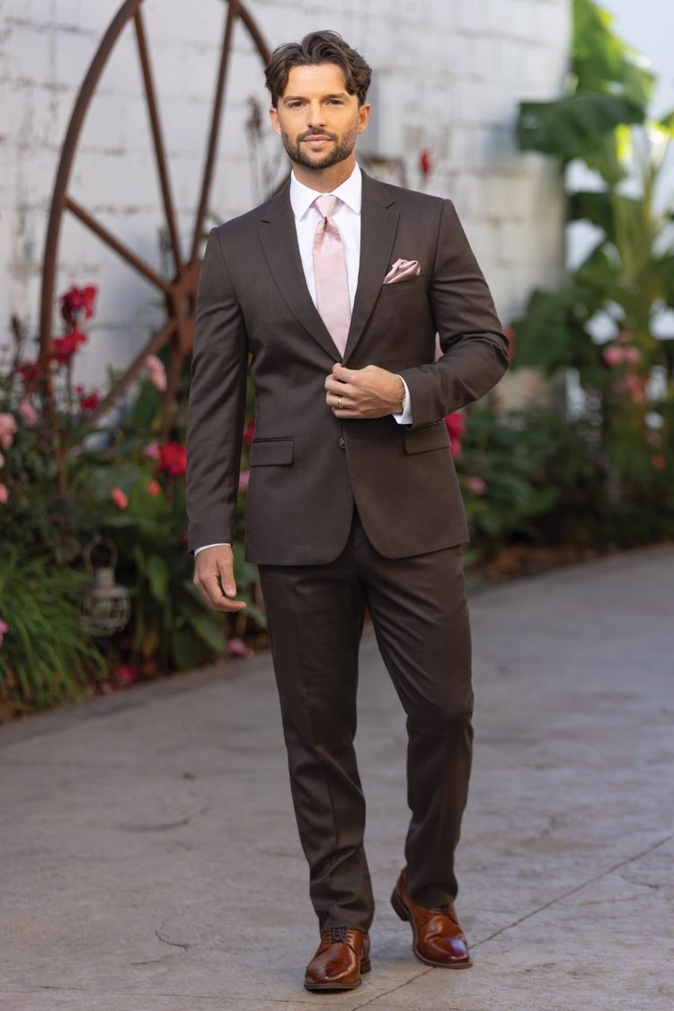 man in dark brown stretch suit coat, pant, and vest coupled with a white dress shirt and light pink windsor tie in a garden courtyard